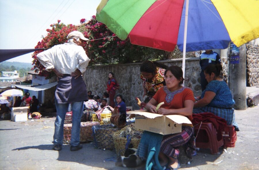 A travel photo of a woman sitting under a colourful umbrella on a Guatemalan street corner