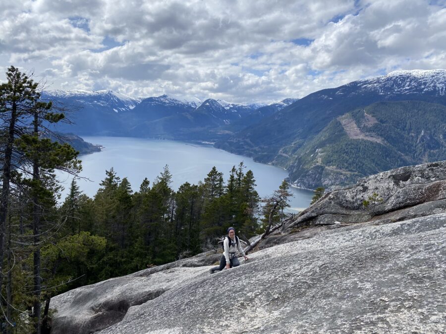 A girl leaning on her knees on a steep rock face, with the ocean in the background. Part of the Stawamus Chief in Squamish, and a perfect stop for a BC summer road trip!