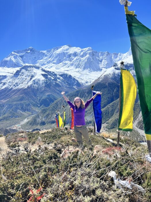 A girl standing with her arms up with snowy mountains behinds her and large, colourful, prayer flags near by.