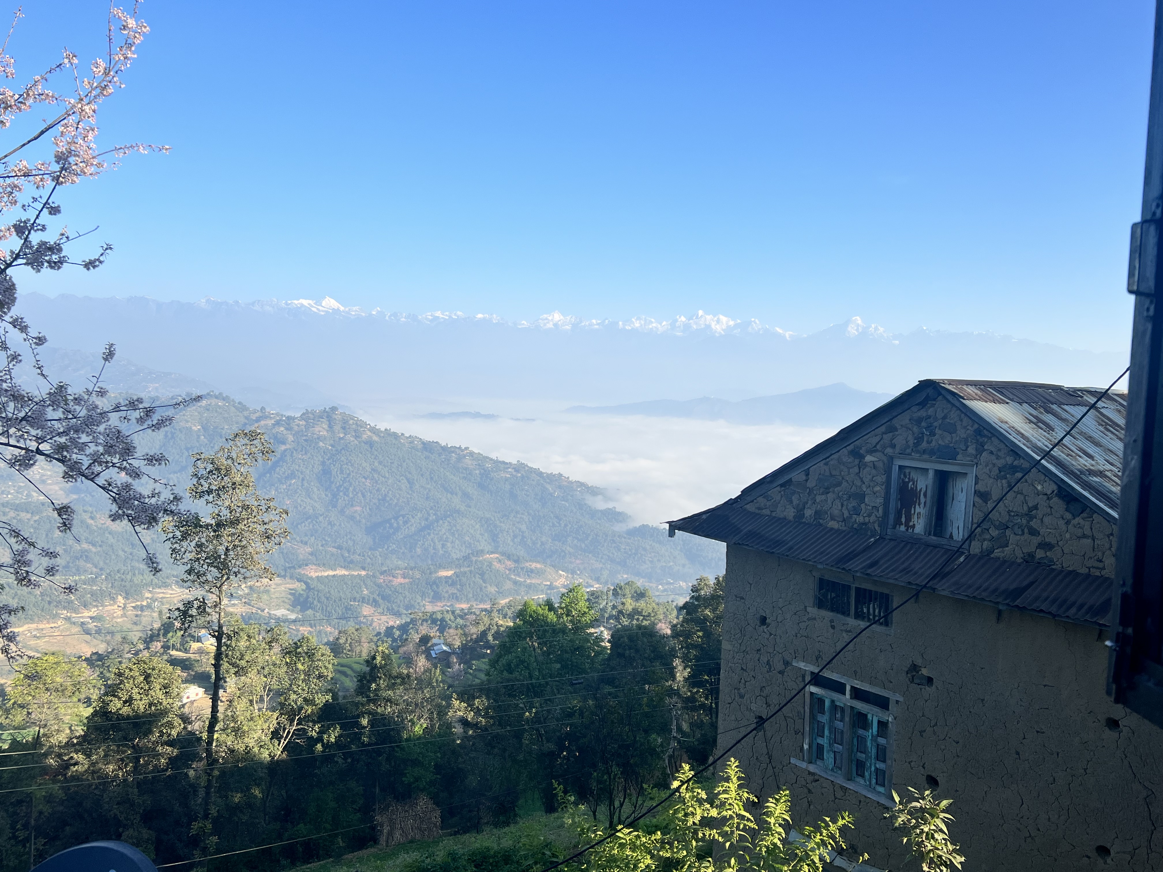 A house with greenery in the foreground and mountains in the background, in Patlekhet Nepal.