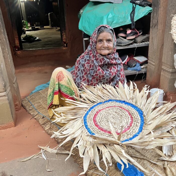 An elderly Nepali woman sitting on a mat on the ground, posing with a beautiful mat she is in the process of making.