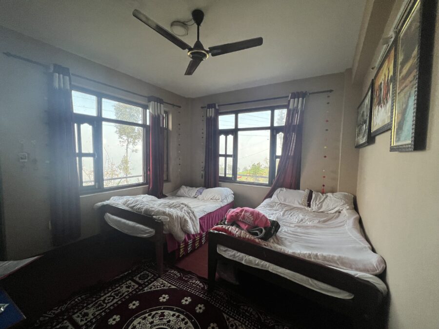 A room with two beds, and two windows with red curtains, that is the accommodation for a Nepali homestay. 