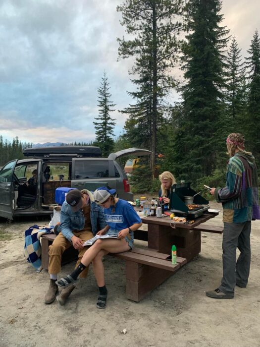 A group of people gathered around a picnic table, with a camping stove set up on it and some people cooking. 