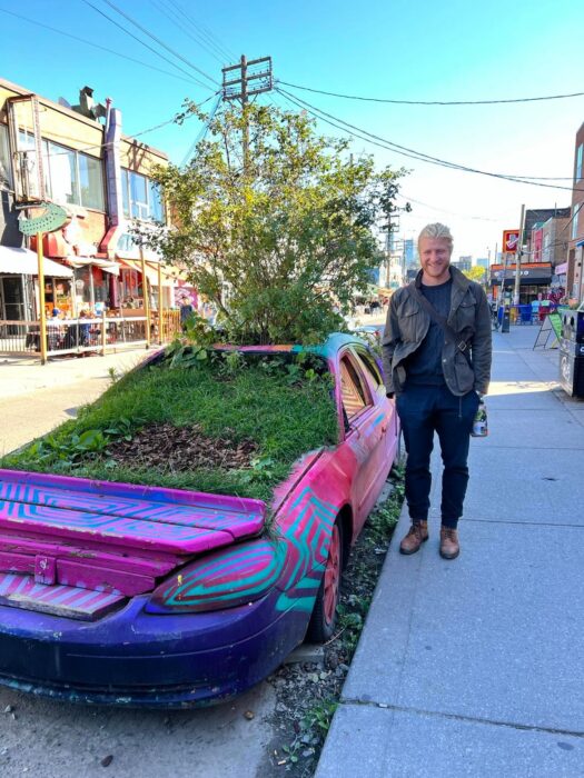 A man standing next to a pink car with grass and trees growing out of it, in Toronto, ON, Canada.