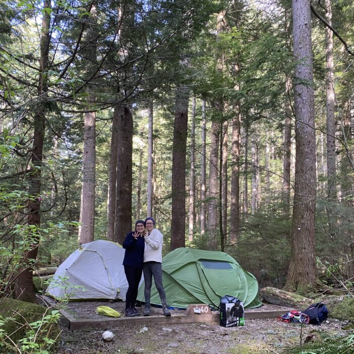 Two girls posing in front of some tents at a camp spot, a great way to save money while road tripping in Canada.