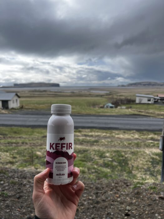 A bottle of Kefir being held up outside in Iceland with a road, grass, houses, and the ocean visible in the background. 