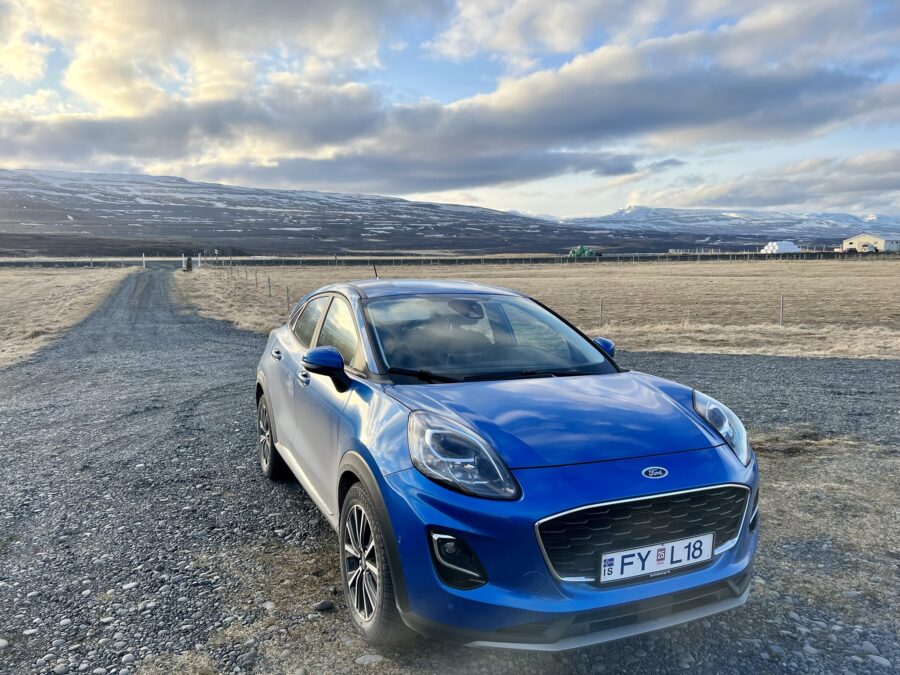 A blue car parked at the end of a driveway with mountains in the background.