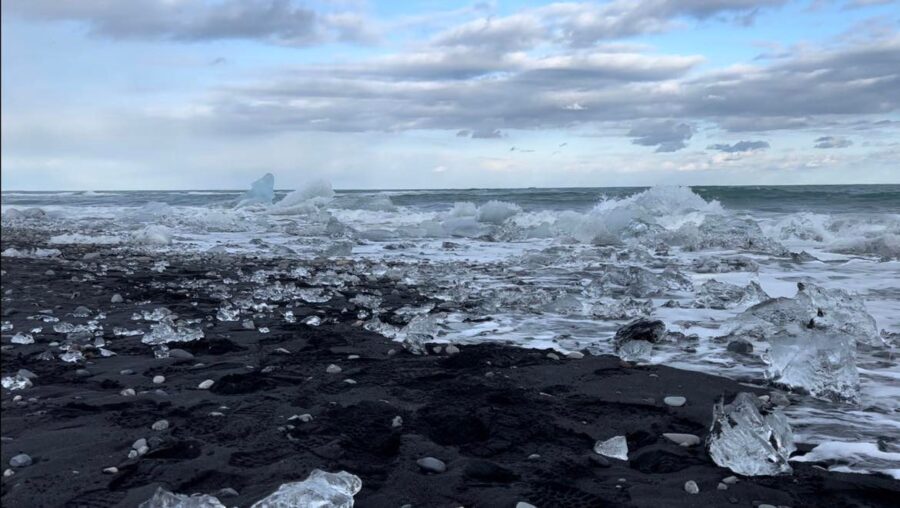 An image of a black sand beach where it meets the ocean, covered in chunks of ice, and clouds in the sky. 