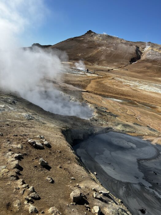 In the foreground there is some dark mud, and a hole with steam coming out. In the background there is a barren hill and you can see another spot with steam coming out. 