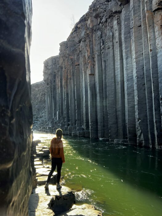 A woman standing in a canyon of hexagonal pillars, Stuðlagil Canyon in Iceland, beside a river where the water looks green. 