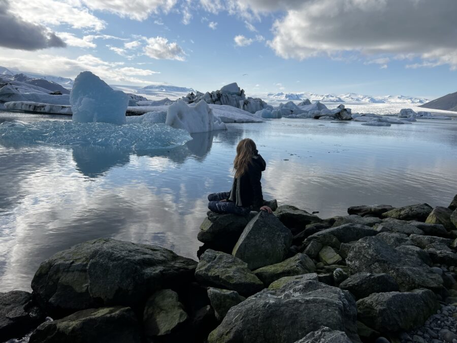 A woman sitting on a rock looking out over Jökulsárlón lagoon in Iceland with large chunks of ice floating in it.