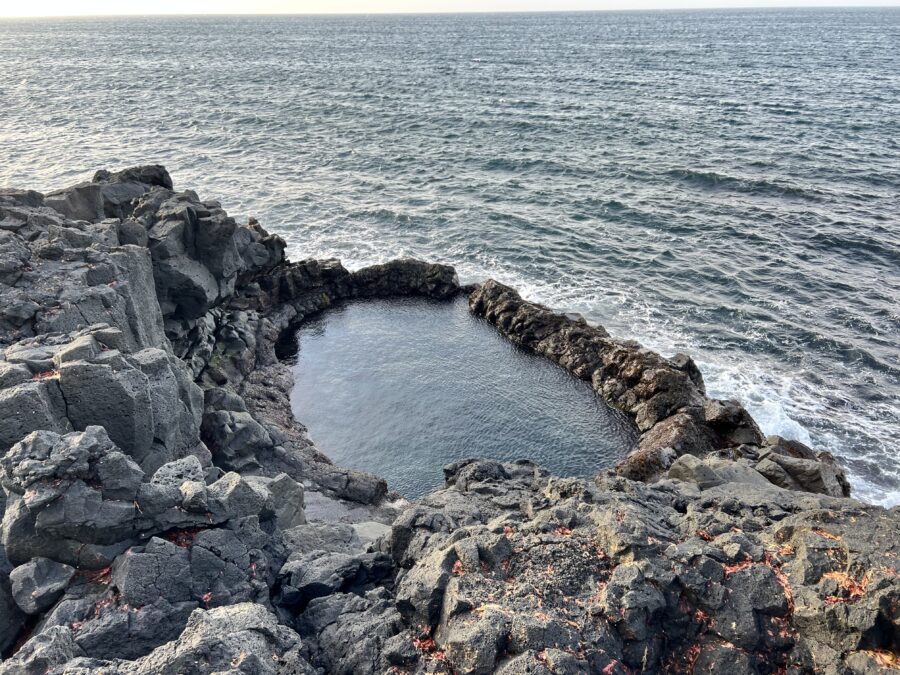 A rocky cliff edge beside the ocean, with a rock formation that resembles a swimming pool filled with water.