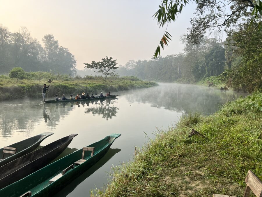 A wide river with 3 parked, long, boats in the foreground. In the middle of the river is another long boat full of people sitting in it with one person standing on the back holding a long stick to push the boat along. The banks on either side of the river are grassy.