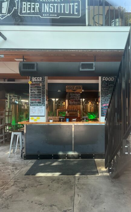 The inside of the Kelowna Beer Institute. In the photo you can see a counter with the sign "Order Here" about it and chalkboard with beer and food menus on either side. 