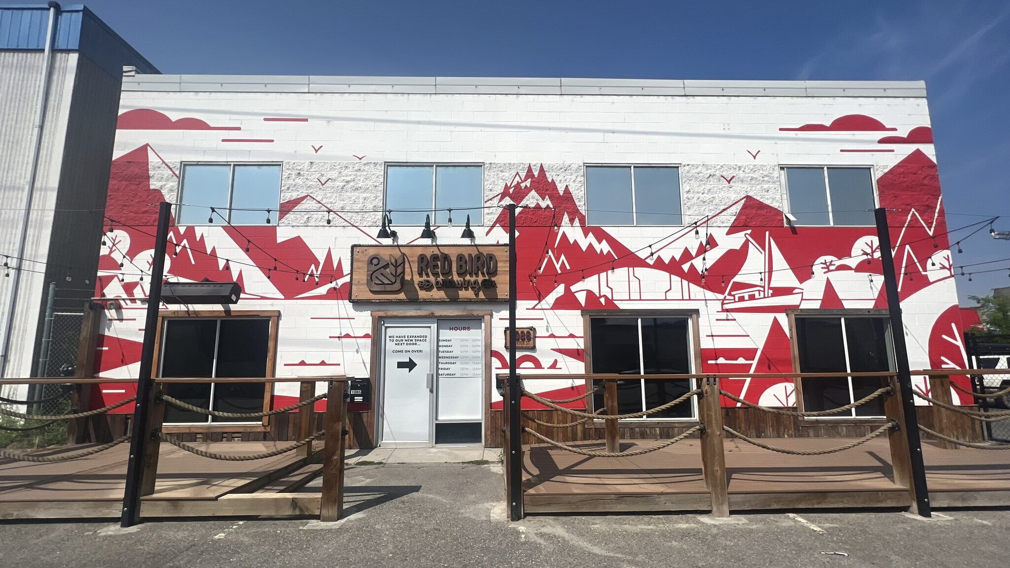 The front of Red Bird Brewing in Kelowna. The building is white with a painting of mountains, a lake, and a boat, all in red. 