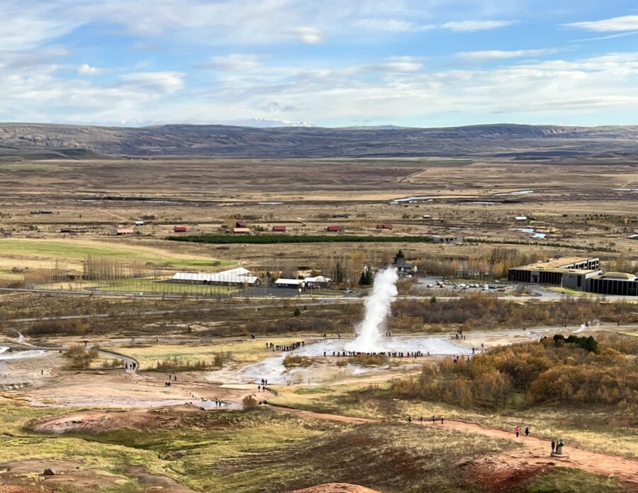 A far away photo of a geysir erupting surrounded by fields and flatland with some hills in the background. 