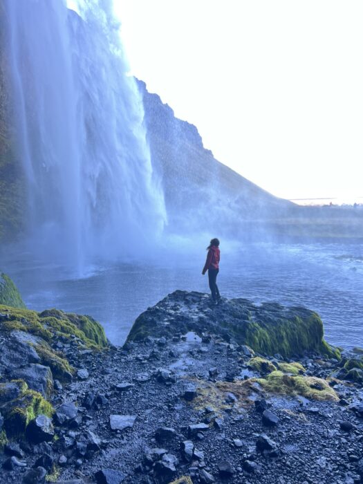 A girl in a red jacket looks up at a huge waterfall. 