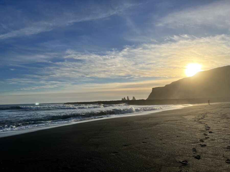 Waves rolling up on a black sand beach in Vik Iceland with footprints in the sand. The sun is starting to set behind a cliff in the background. 