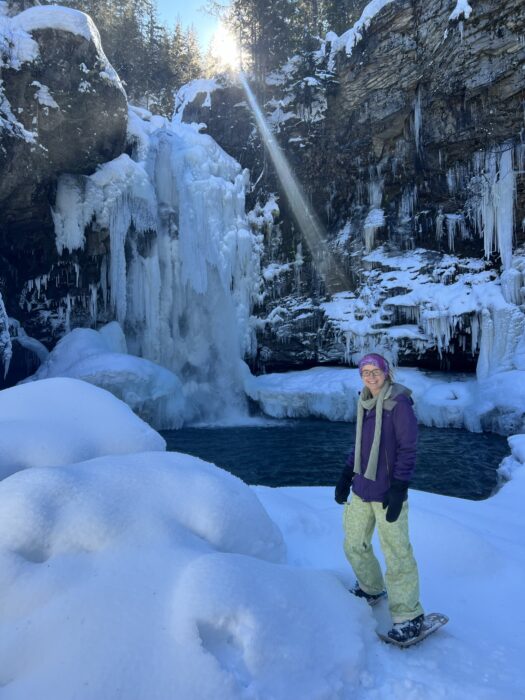 A girl standing in front of a frozen Sutherland Falls with snowshoes on. 