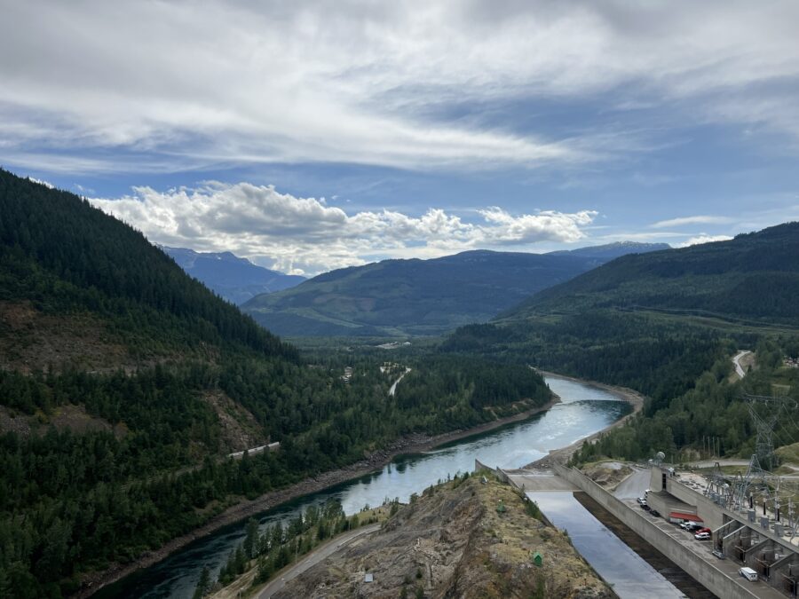 A beautiful view of a winding blue river (the Columbia), mountains covered in green trees, and a partially cloudy blue sky, with the spillway of the Revelstoke Dam visible in the corner of the picture