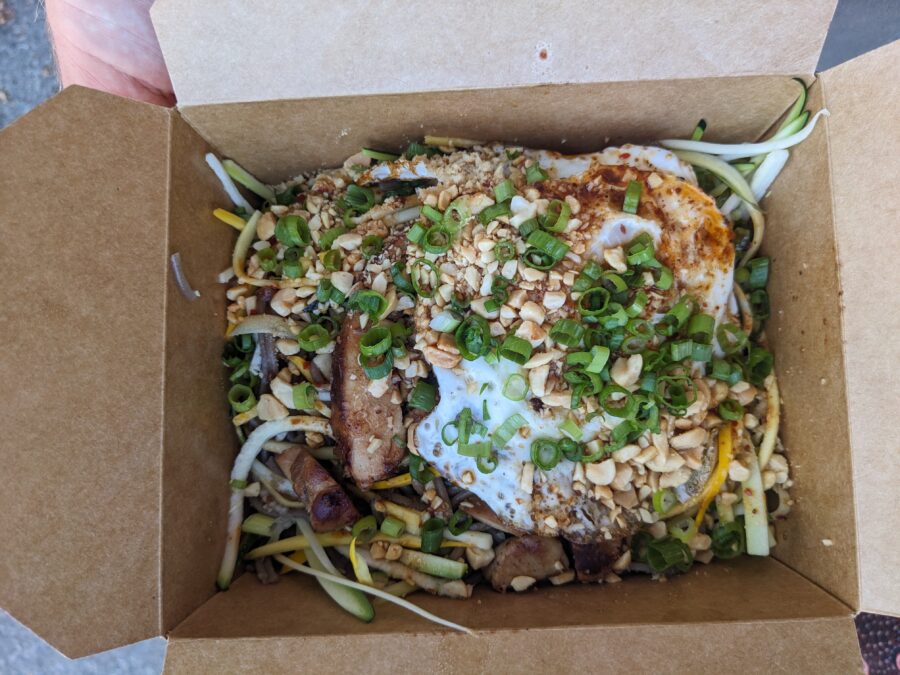 A noodle dish with chicken, peanuts, egg, and green onion in a takeout box from The Lucky Pup