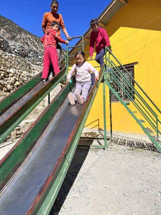 A double metal slide in Nepal with two kids sliding down one side and two other kids standing on the other side