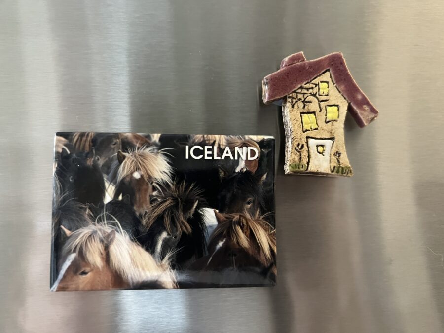 Two magnets on a metallic fridge, one with horses that says "Iceland" and another handpainted and in the shape of a little house