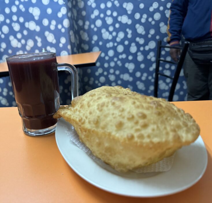 A glass of api, a hot Peruvian drink made of purple corn beside a large empanada it is served with