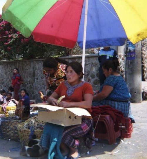 A travel photo of a woman sitting under a colourful umbrella on a Guatemalan street corner
