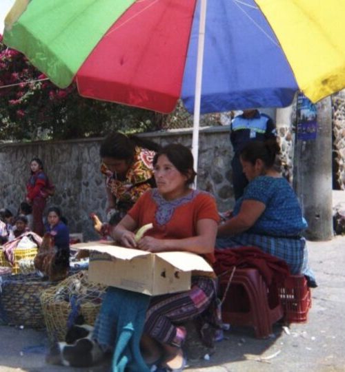 A travel photo of a woman sitting under a colourful umbrella on a Guatemalan street corner